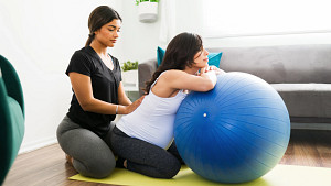 Mujer embarazada apoyada en una pelota de ejercicio con un entrenador que ayuda a estirar la parte inferior de la espalda.